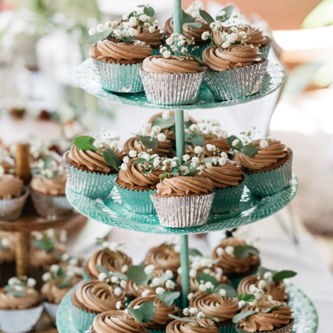 Cupcakes mit Schokoladenglasur und kleinen weißen Blumen sind auf einem dreistöckigen grünen Ständer angeordnet. Die Cupcakes sind mit grünen Blättern und weißen Blumen geschmückt und stehen auf einem hellen Tisch.