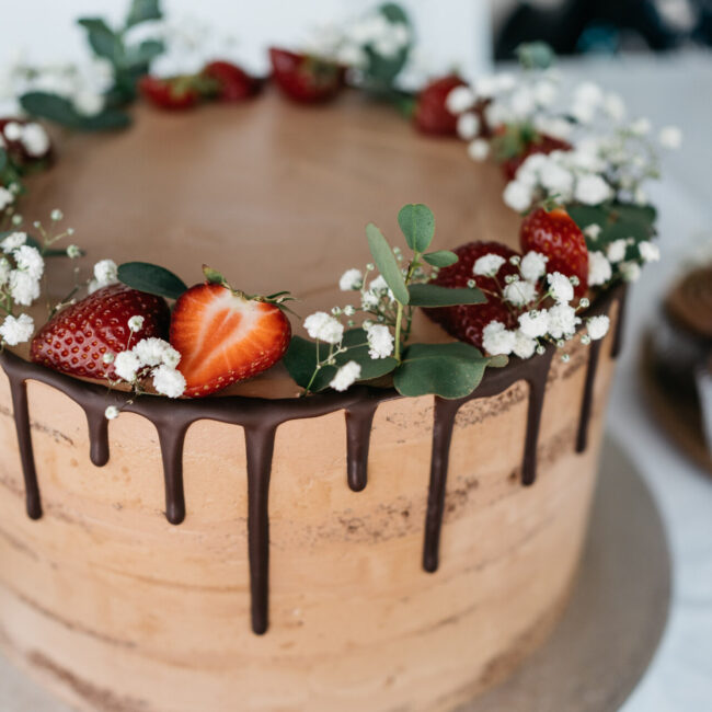Ein Schokoladenkuchen mit glatter, glasierter Oberfläche und Schokoladenglasur, die an den Seiten heruntertropft. Die Oberseite ist mit frischen Erdbeeren, grünen Blättern und weißen Blumen dekoriert, die kreisförmig um den Rand angeordnet sind.