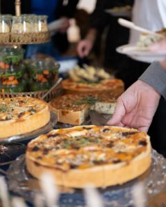 Die Hand einer Person serviert ein Stück Gemüsequiche von einem Buffettisch. Der Tisch ist mit verschiedenen ganzen Quiches und Salaten in Glasbehältern gefüllt. Die Szene lässt auf eine zwanglose Essensatmosphäre schließen.