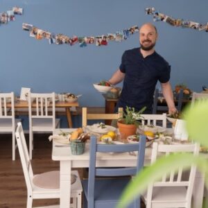 Eine lächelnde Person mit Bart steht in einem Essbereich und hält eine Schüssel Salat in der Hand. Der Raum hat blaue Wände, die mit einer Reihe von Fotos dekoriert sind. Mehrere Tische sind mit Tellern voller Essen und Pflanzen gedeckt. Weiße und blaue Stühle umgeben die Tische.