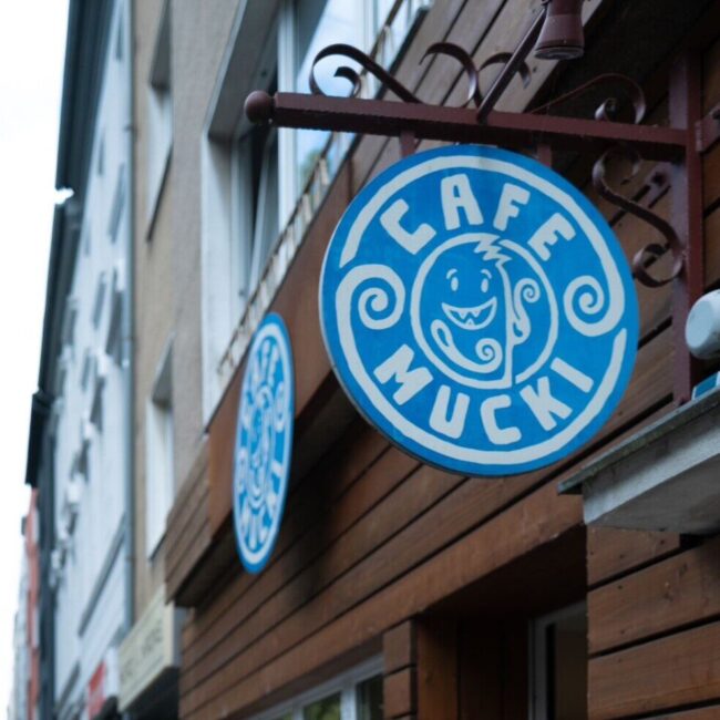 Das Bild zeigt ein Hängeschild für „Cafe Mucki“ mit einem stilisierten Gesichtslogo in Blau und Weiß. Das Café liegt an einer Straße mit ähnlichen Schildern auf beiden Seiten, angebracht an einer Holzfassade in einer städtischen Umgebung.