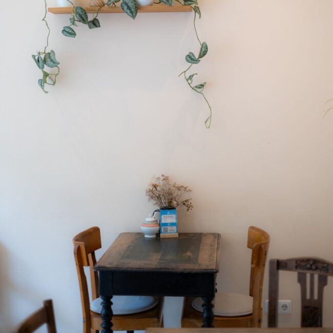 Ein kleiner Holztisch mit zwei Stühlen steht an einer schlichten Wand. Darüber befindet sich ein Holzregal mit einigen Dekoartikeln und einer Pflanze mit Ranken. Auf dem Tisch steht eine kleine Vase mit Trockenblumen.