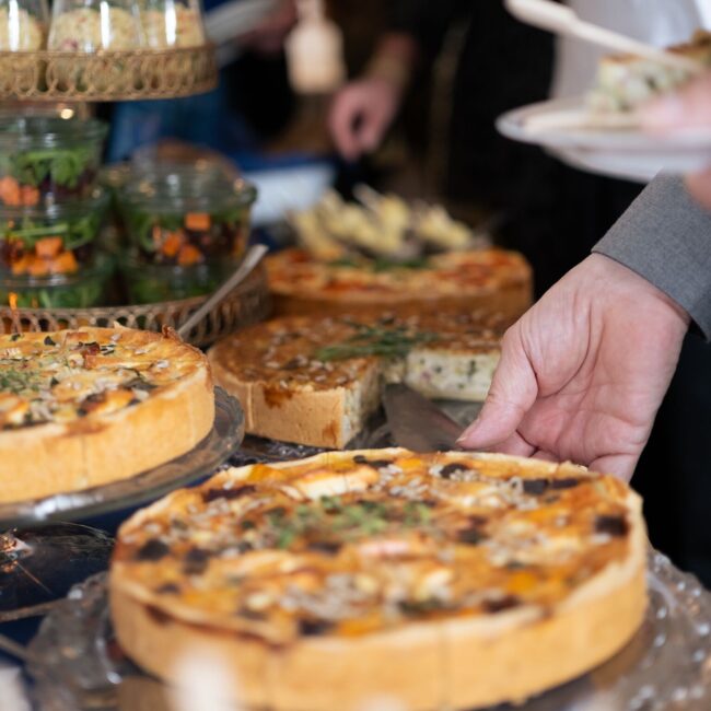 Eine Nahaufnahme mehrerer frisch gebackener Quiches auf einem Tisch. Die Hand einer Person serviert ein Stück mit einem Spatel. Im Hintergrund sind kleine Salate in Behältern und andere Gerichte zu sehen.