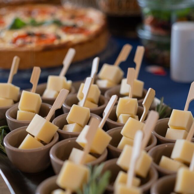 Kleine Schüsseln mit Käsewürfeln auf Spießen sind auf einem Tablett angeordnet und mit Rosmarin garniert. Der unscharfe Hintergrund zeigt Pizzas und Salatschüsseln, wodurch eine Buffet-ähnliche Atmosphäre entsteht.