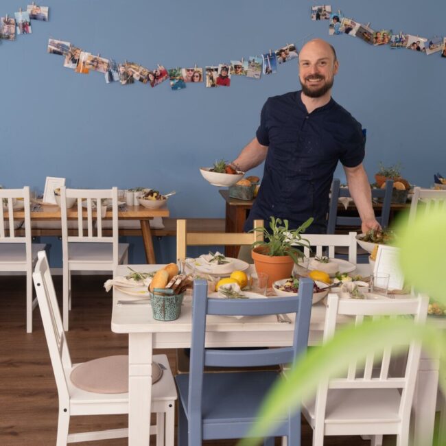 Ein Mann mit Bart steht lächelnd in einem Essbereich und hält Teller mit Essen in der Hand. Der Raum hat eine blaue Wand mit einer Fotogirlande. Weiße Stühle umgeben einen mit Pflanzen, Brot und Zitronen gedeckten Tisch. Im Vordergrund steht eine grüne Pflanze.