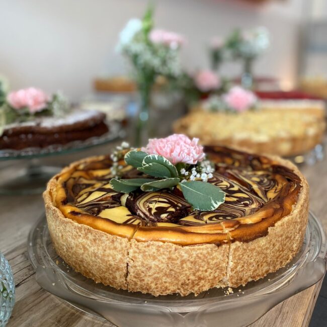 Ein marmorierter Käsekuchen, garniert mit rosa Blumen und Grünpflanzen, präsentiert auf einem Tortenständer aus Glas. Der Tisch ist mit weiteren Blumenarrangements und verschwommenen Torten im Hintergrund geschmückt.