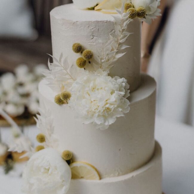 Eine dreistöckige weiße Hochzeitstorte, geschmückt mit weißen Blumen, Zitronenscheiben und kleinen gelben Pompon-ähnlichen Verzierungen. Die Torte steht auf einem goldenen Untersatz und im Hintergrund steht ein Tisch mit ähnlicher Blumendekoration.