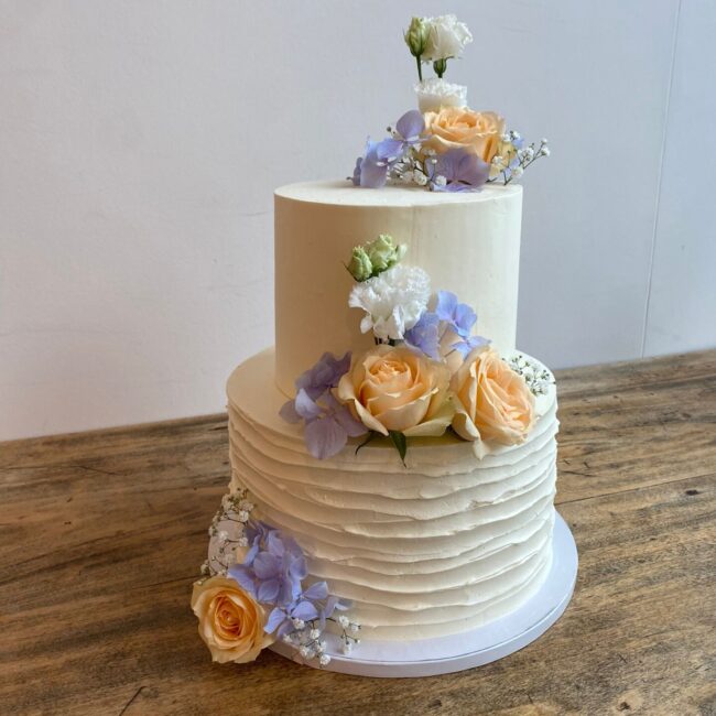 Eine zweistöckige weiße Torte, dekoriert mit orangefarbenen Rosen, weißen und violetten Blumen und Schleierkraut auf einem Holztisch. Die untere Etage der Torte weist ein strukturiertes, gerüschtes Muster auf.
