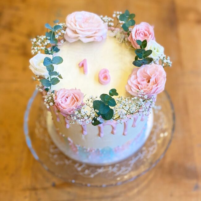 Ein runder, weißer Kuchen mit rosafarbener Glasur, verziert mit rosafarbenen und weißen Rosen, Eukalyptusblättern und kleinen weißen Blumen. Oben ist die Zahl „16“ in Rosa platziert. Der Kuchen steht auf einem Glasständer über einem Holztisch.
