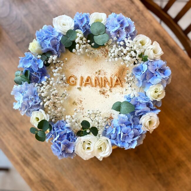 Ein runder Kuchen, verziert mit einer Vielzahl blauer und weißer Blumen, darunter Hortensien und Rosen, akzentuiert mit Grünpflanzen. Das Wort „GIANNINA“ ist in goldenen Buchstaben auf der cremefarbenen Oberfläche des Kuchens geschrieben, der auf einem Holztisch steht.