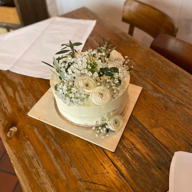 Eine einstöckige weiße Torte, geschmückt mit weißen Rosen und grünen Blättern, steht auf einem Holztisch, daneben liegt eine weiße Tischdecke.