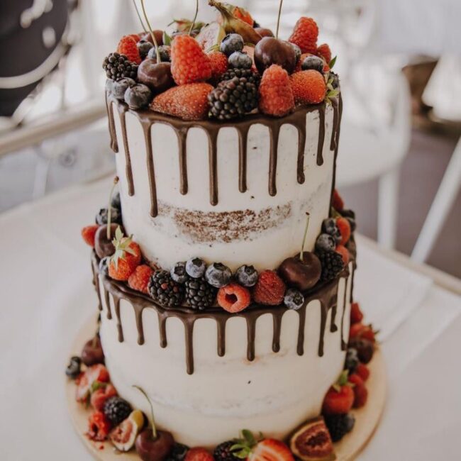 Ein zweistöckiger Naked Cake, dekoriert mit frischen Beeren und Kirschen. Schokoladenganache tropft an den Seiten herunter und sorgt für einen satten Kontrast zum hellen Zuckerguss. Der Boden ist mit weiteren Früchten geschmückt, wodurch eine lebendige und appetitliche Präsentation entsteht.