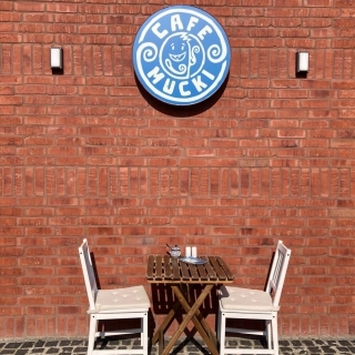 Ein kleines Straßencafé mit zwei weißen Stühlen und einem Holztisch vor einer roten Backsteinwand. Darüber hängt ein blaues rundes Schild mit einem verspielten Design und dem Text „Cafe Mucki“.